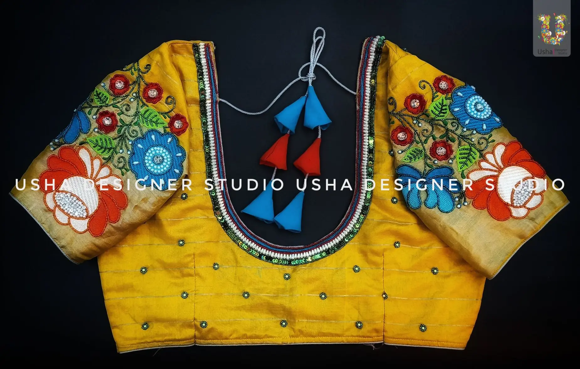 A vibrant yellow bridal blouse from Usha Designer Studio with heavy, multi-colored floral Aari work embroidery on the sleeves in shades of blue, red, and orange. The U-shaped neckline is embellished with a beaded border, and the blouse features matching tassels.