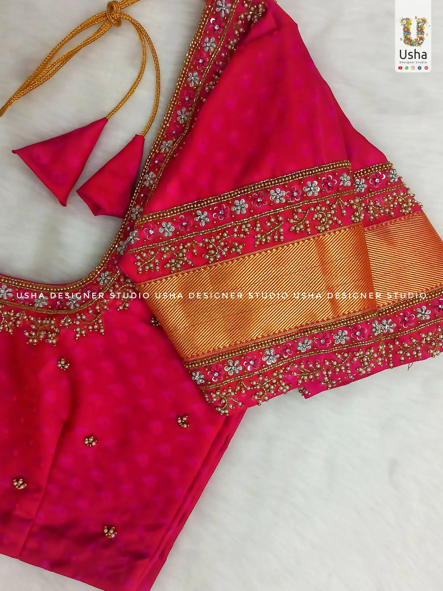 bright rani pink bridal blouse from Usha Designer Studio. The elbow-length sleeves feature a wide golden zari border, framed by intricate Aari and Maggam bead and stone work. Similar embroidery adorns the neckline.