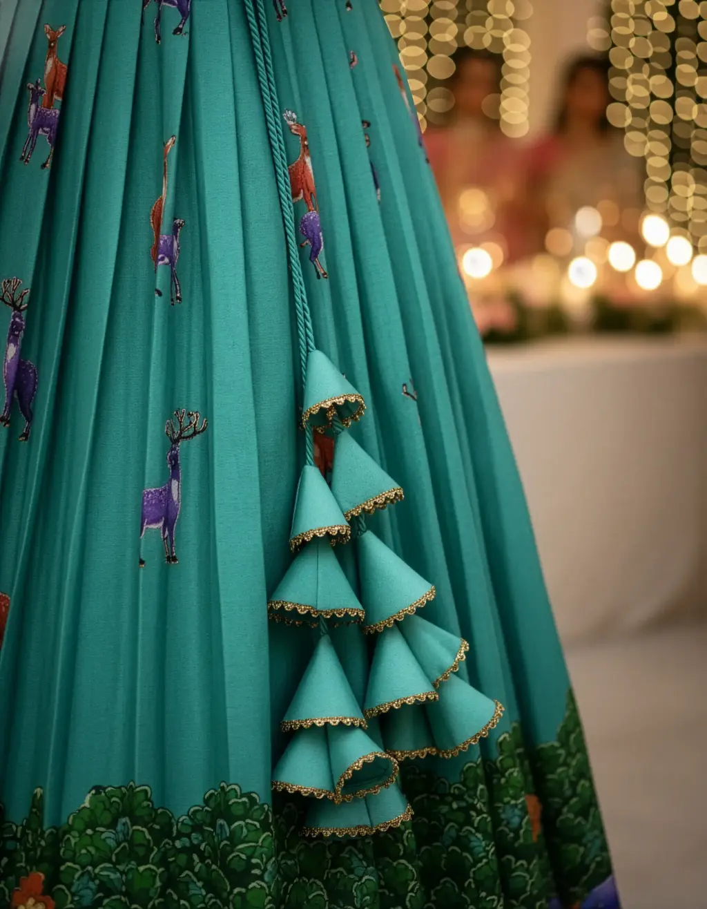 Image of a tailor-made teal-blue crop top lehenga featuring a multi-colored forest/jungle animal print, long sleeves, V-neck Aari scallop, and heavy triangular latkans on the pleated skirt.