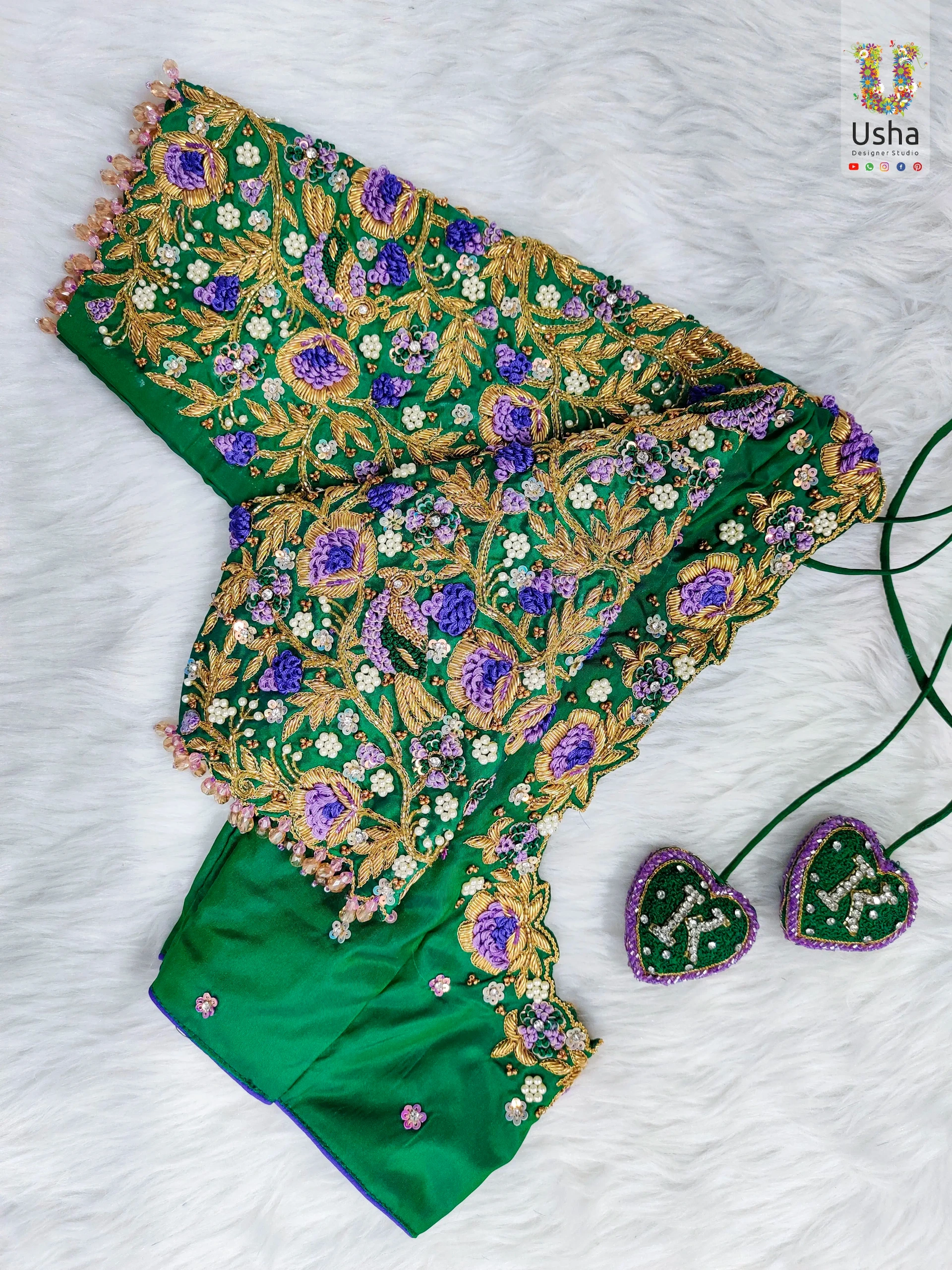 Tailor-Made Green Floral Aari Embroidery Blouse | Usha Designer Studio Vijayawada Tailor-made green blouse with dense floral Aari embroidery in gold, purple, and white, beaded sleeve edges, and matching personalized heart tassels, designed by Usha Designer Studio, Vijayawada, displayed on a white fur background.