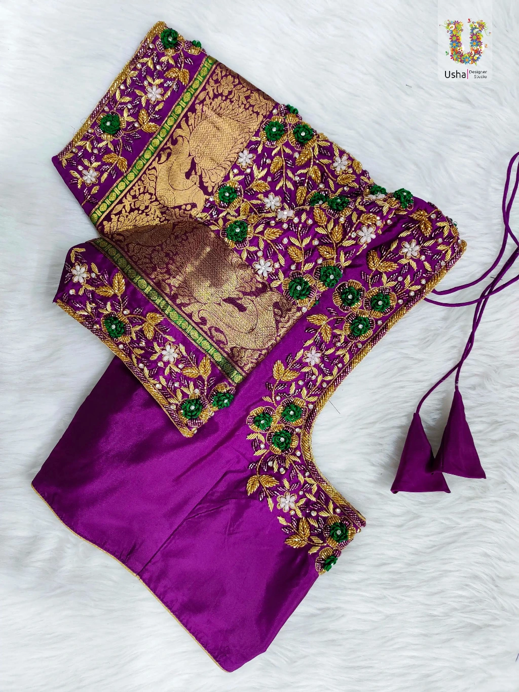 Custom purple silk designer blouse by Usha Designer Studio featuring heavy gold Aari and Zardosi embroidery, emerald green beadwork, pearl detailing on the neckline, and a traditional gold woven zari border on the sleeves.