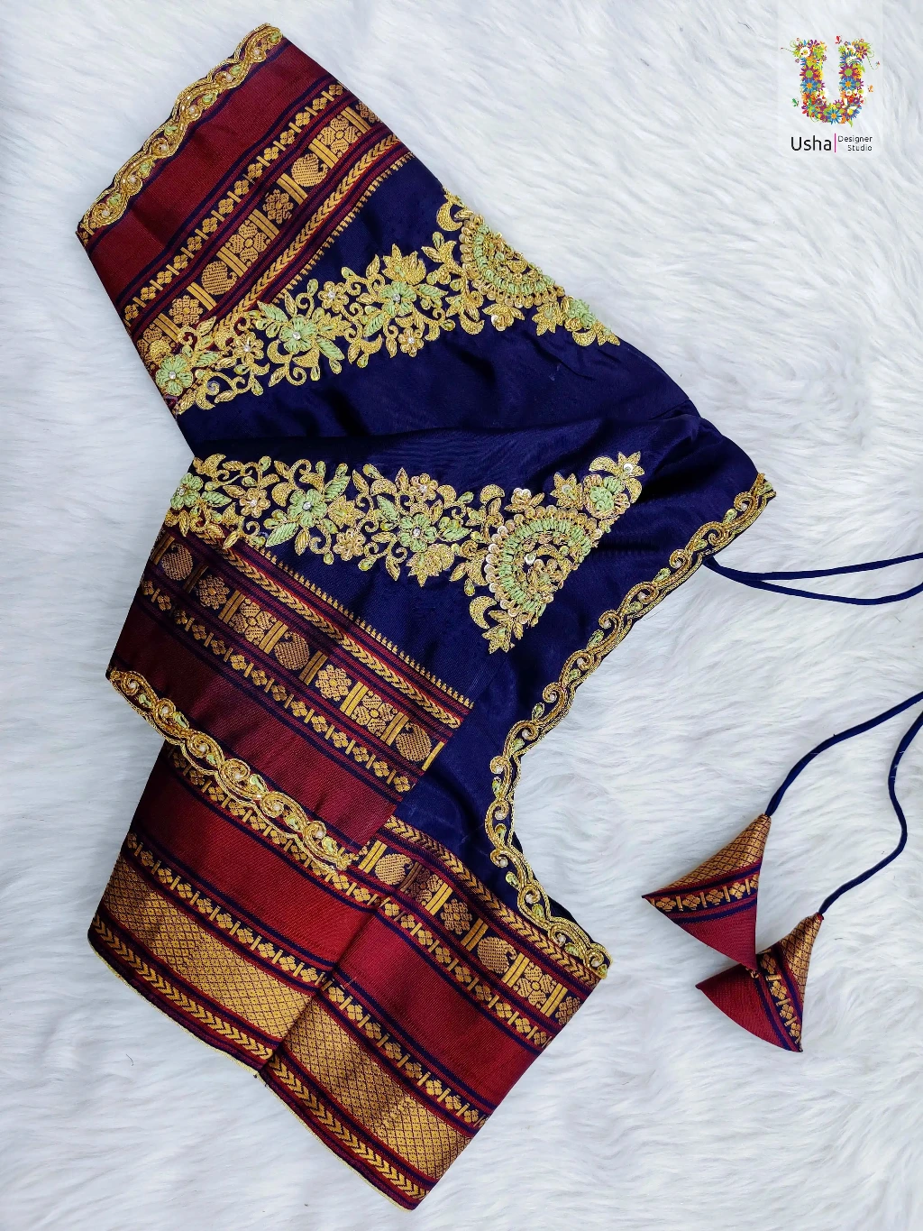 Navy blue silk designer blouse by Usha Designer Studio featuring intricate Aari embroidery, heavy gold floral zardosi, maroon Kanjeevaram style borders, scalloped edges, and matching fabric tassels on a white background.