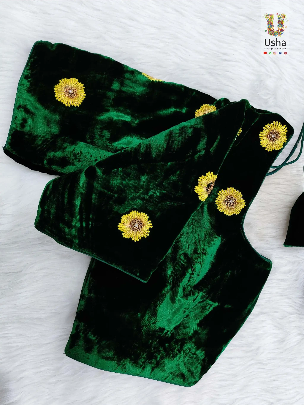 Emerald green crushed velvet designer blouse pieces featuring bright yellow hand-embroidered floral patterns with detailed centers, laid out on a white fur background with the Usha Designer Studio logo.