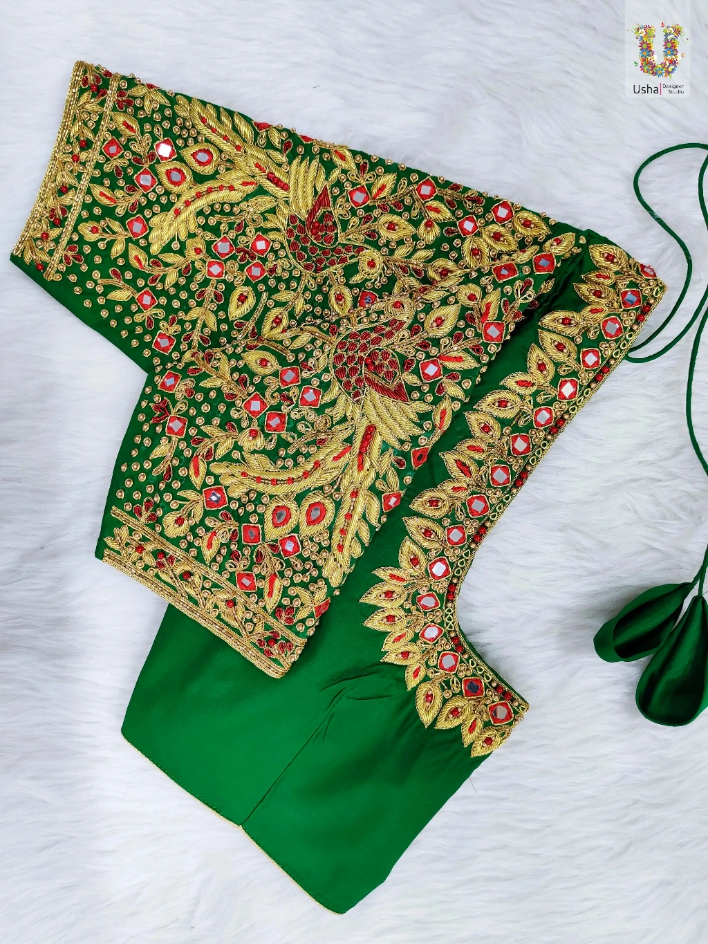 Usha Designer Studio emerald green bridal blouse piece featuring heavy hand-crafted gold Aari embroidery, red bead accents, peacock and floral motifs on sleeves, diamond mirror work, and a scalloped neckline border on a white fur background.